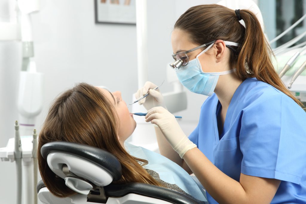 Dentist wearing eyeglasses gloves and mask examining a patient teeth with a dental probe and a mirror in a clinic box with equipment in the background
